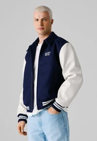 Młody mężczyzna z krótkimi platynowymi włosami, ubrany w granatowo-białą kurtkę Red Bull Racing oraz jasnoniebieskie jeansy, stojący na tle szarego tła.