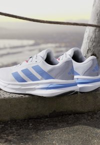 Des chaussures de sport grises avec des accents bleus, présentant une texture en maille, un logo à trois bandes bien en vue et un rembourrage Cloudfoam, posées sur une surface en béton.