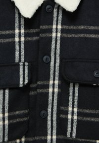 Giacca a maniche lunghe a quadri in bianco e nero con due tasche a pattina e colletto in sherpa bianco, visibile in primo piano.
