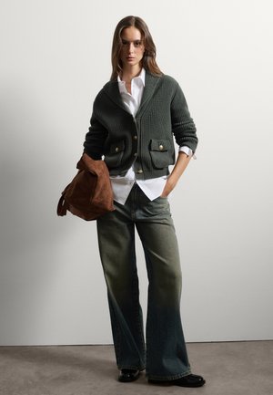 Olivgrüner Strickcardigan mit gerippter Textur und goldenen Knöpfen, kombiniert mit einem weißen Hemd, dazu eine flared Jeans und eine braune Wildledertasche.