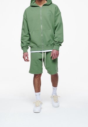 Groene zip-hoodie met een voorzak en bijpassende shorts. Gemaakt van zachte stof. Kenmerkt een trekkoord en witte sportkousen.