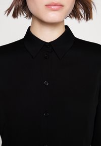 Camicetta nera con colletto classico, texture del tessuto liscia e bottoni neri lungo la patta anteriore.
