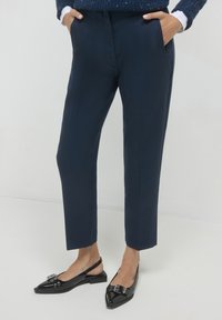 Pantaloni blu navy con una vestibilità su misura, dotati di tasche laterali e una texture liscia. Abbinati a scarpe slingback nere lucide con decorazioni.