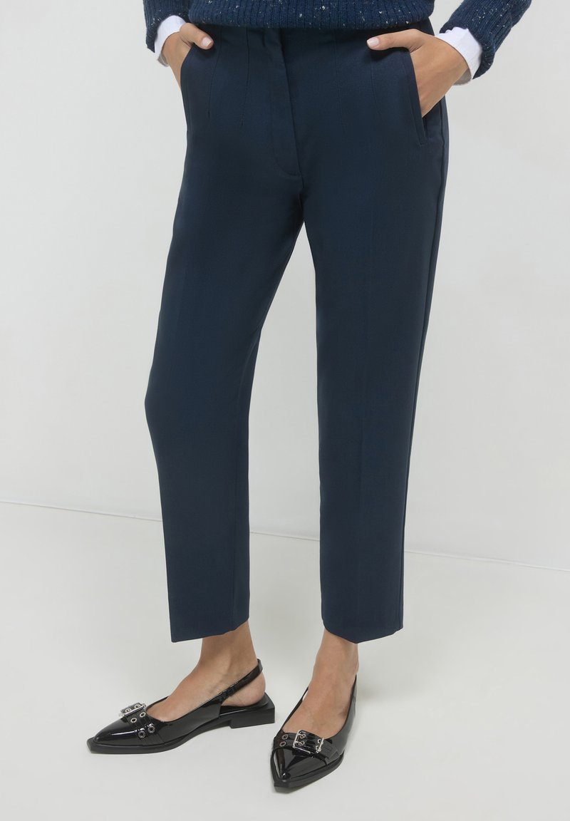 Pantaloni blu navy con una vestibilità su misura, dotati di tasche laterali e una texture liscia. Abbinati a scarpe slingback nere lucide con decorazioni.