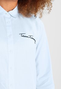 Light blue button-up shirt with a pointed collar, featuring black embroidered "Team Tommy" text on the left side. Smooth fabric texture.