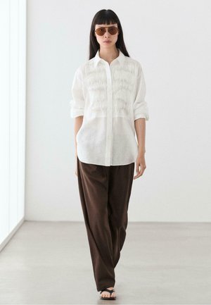 Woman walking indoors wearing brown pants, white textured button-up shirt, dark sunglasses, and black sandals on a light floor and wall background.