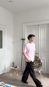 Άτομο που φοράει ένα ανοιχτό ροζ μπλουζάκι και μαύρο παντελόνι περνάει μπροστά από μια λευκή πόρτα. Ο χώρος διαθέτει ξύλινο δάπεδο, μια καρεκλάκι με μοτίβο και μοντέρνα διακόσμηση.