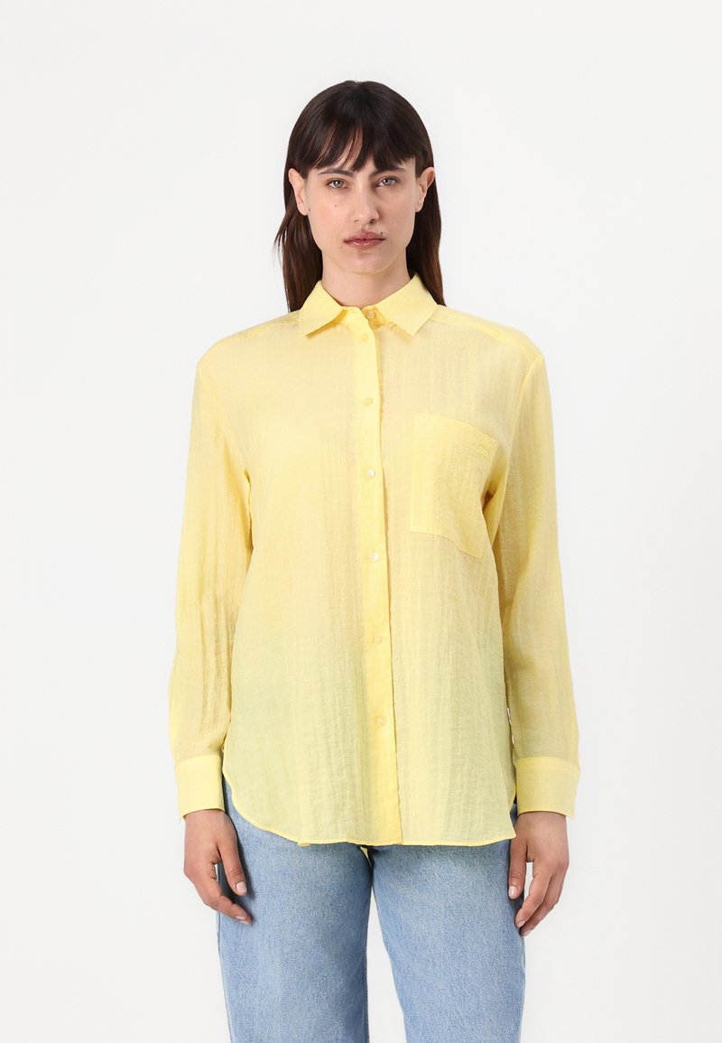 Camicia gialla con bottoni, in tessuto leggero e stropicciato, con una tasca sul petto, maniche lunghe e orlo arrotondato. Abbinata a jeans azzurri chiari.