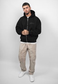 Sort zip-up hoodie med logo, beige cargo bukser og hvide sneakers. Har en afslappet pasform og hætte, med synlige søm detaljer.