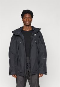 Casaco impermeável preto com capuz, apresentando bolsos com fecho éclair e um logótipo no peito. O design é descontraído e funcional.