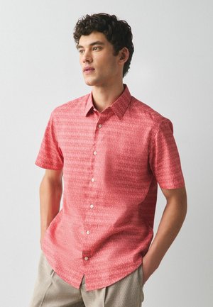 Mężczyzna z kręconymi włosami, ubrany w koralową czerwoną, krótko rękawową koszulę z subtelnym wzorem fal, beżowe spodnie, stoi z rękami w kieszeniach.