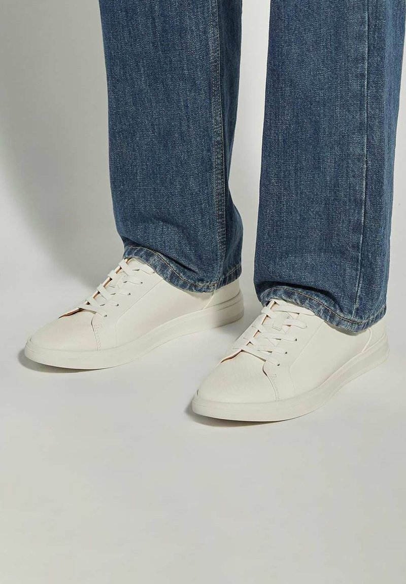 Baskets blanches en matériau texturé, bout arrondi, semelle blanche plate et lacets ton sur ton, associées à un jean en denim bleu.