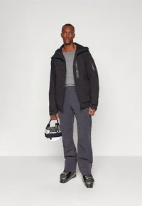 Jaqueta preta isolada com capuz, camada base cinzenta, calças de ski cinza escuro e botas de ski pretas, segurando um capacete branco com detalhes pretos.