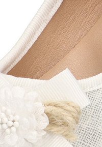Zapato textil blanco con superficie texturizada, flor decorativa blanca con cuentas y un acento de cuerda natural cerca del área del dedo del pie.