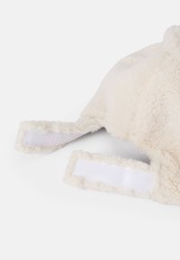 Bonnet en polaire couleur crème avec une texture douce, comportant des sangles en Velcro blanches sur les côtés pour le maintenir. Design arrondi pour un ajustement confortable.