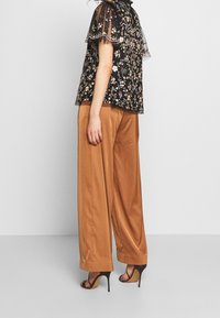 Sort blomstret top med gennemsigtige, voluminøse ærmer parret med højtaljede, løstsiddende brune bukser og sorte højhælede sandaler.