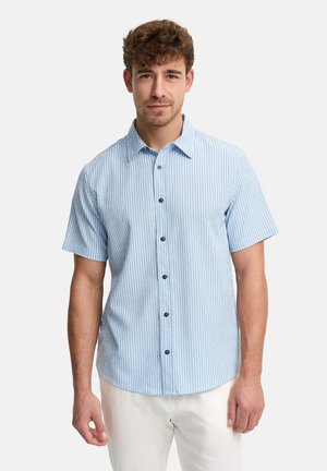 Mężczyzna ubrany w jasnoniebieską i białą koszulę na krótki rękaw z pionowymi paskami oraz białe spodnie na jednolitym tle.