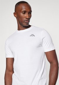 White short-sleeve t-shirt made of cotton, featuring a black Kappa logo stitched on the left chest area. Smooth texture, classic fit.
