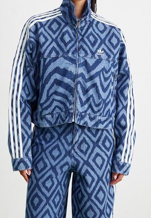 Woman wearing a blue patterned Adidas track jacket and matching pants with white stripes on sleeves and pant sides.