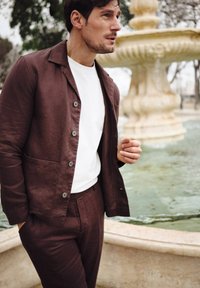 Brun linnejacka med en vit skjorta under, med knappar och sidofickor; står nära en fontän i en utomhusmiljö.