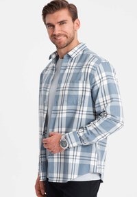 Blauwe en witte geruite shirt gemaakt van katoen, met een knoopsluiting aan de voorkant, een borstzak en lange mouwen. Gdragen over een grijs T-shirt.