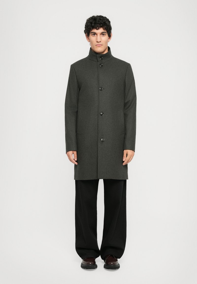 Manteau en laine gris foncé avec un col haut, fermeture à boutons à l'avant, et deux poches latérales, porté sur un pantalon noir ample et des chaussures sombres.