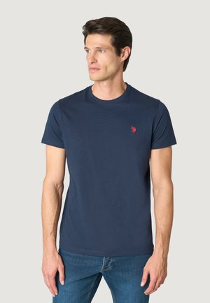 Uomo che indossa una t-shirt blu navy a maniche corte con scollo rotondo e un piccolo logo rosso sul petto sinistro, abbinata a un paio di jeans blu.
