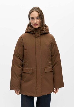 Femme portant une veste matelassée marron à capuche avec poches à rabat, debout devant un fond blanc uni.