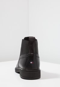 Musta nahkasaapas nilkkapituisella, pyöreällä kärjellä, sileällä pinnalla, vähäisellä ompeleella ja pienellä logolla sivussa.