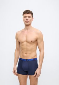 Άνδρας χωρίς πουκάμισο, όρθιος, φορώντας μπλε σκούρα μποξεράκια Puma, μπροστά σε ένα απλό φωτεινό φόντο, κοιτάζοντας ευθεία με ουδέτερη έκφραση.