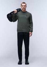 Maglione texturizzato verde oliva con collo rotondo, abbinato a pantaloni neri affusolati e scarpe nere. Il modello porta una borsa a tracolla nera.