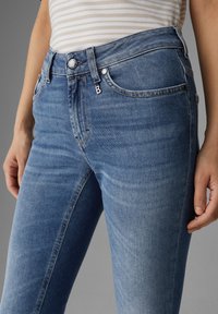Femme portant un jean en denim bleu ajusté et un haut beige à rayures, se tenant avec les mains détendues sur les côtés devant un fond gris.