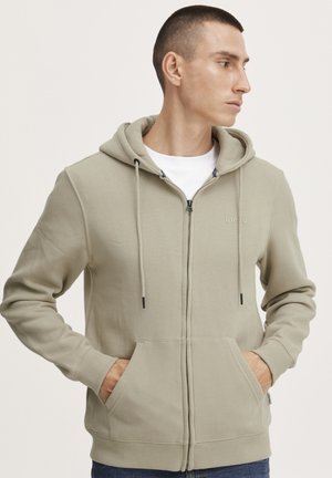 Sudadera con cremallera de color beige claro, con un bolsillo frontal, cordones ajustables y puños acanalados. Presenta un sutil logo de la marca en el pecho.