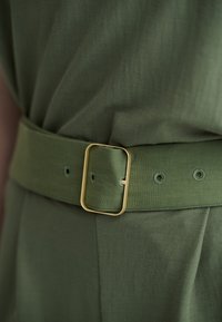 Κοντινό πλάνο σε ζώνη από ύφασμα σκούρου πράσινου ελιάς με ορθογώνια χρυσή αγκράφα, φορεμένη πάνω σε κατάλληλο ρούχο.