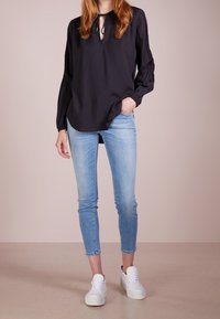 Blouse noire à manches longues avec encolure en trou de serrure, portée avec un jean skinny bleu clair et des baskets blanches sur un fond neutre.