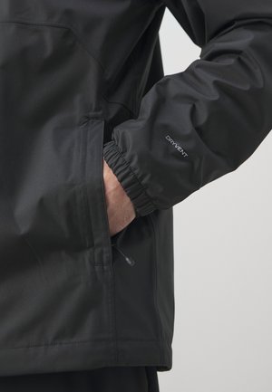 Hand in der Tasche einer schwarzen wasserabweisenden Jacke mit elastischem Bündchen, beschriftet mit „DRYVENT“, auf einem einfarbigen hellen Hintergrund.