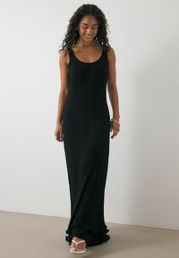 Schwarzes, geripptes Maxikleid mit rundem Ausschnitt, slim fit und volantrigem Saum. Modell trägt Sandalen mit offenen Zehen und steht vor einem schlichten Hintergrund.