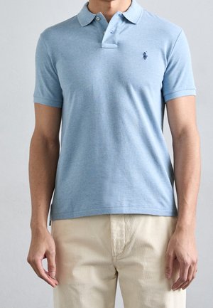 Mann trägt hellblaues kurzärmeliges Poloshirt mit kleinem marineblauem Logo und beigefarbene Cordhose, steht vor einfarbigem Hintergrund.