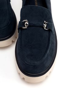 Mocasines de ante azul marino con suela gruesa crema, que presentan un detalle decorativo de hebilla metálica en la parte delantera y un talón ligeramente elevado.