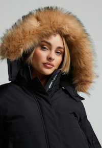 Černá parka s kapucí lemovanou umělou kožešinou. Obsahuje vysoký límec, zipový uzávěr a nenápadné logo. Měkká, texturovaná kožešina v béžové barvě.