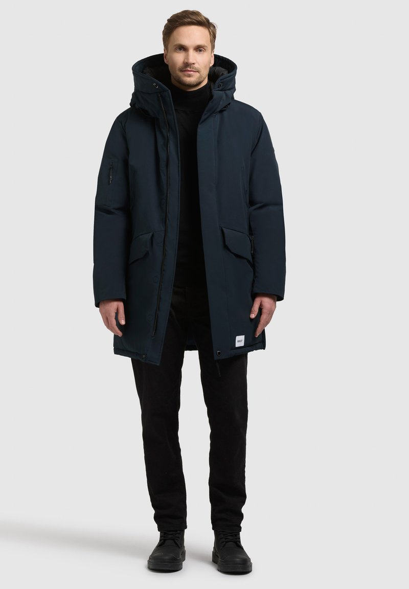 Parka isolante di colore blu navy con cappuccio, dotata di zip frontale, due ampie tasche frontali e un piccolo etichetta del marchio sull'orlo.