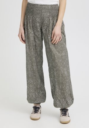 Naine, kes kannab lahtisi, mustrilisi rohelisi pükse, millel on elastsed vöökohad ja sääreotsad, kombineeritud valgete tossudega, mille paelad on mustad.