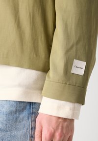 Olijfgroene jas met een gladde textuur, voorzien van een witte corduroy binnenlaag en een Calvin Klein-label op de mouw. Blauwe denim zichtbaar daaronder.