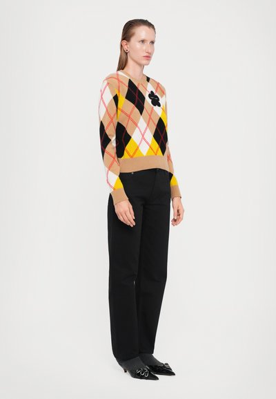 Woman standing wearing an argyle-patterned sweater with black, beige, yellow, and red, paired with black pants and pointed black shoes.