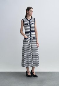 Žena stojí a má na sobě bezrukávé, na knoflíky zapínané šaty s vodorovnými modro-bílými pruhy délky až ke kotníkům a černé boty typu Mary Jane.