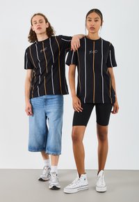 Svarta t-shirts med vita och orange vertikala ränder, bärs med blå denimshorts och svarta cykelshorts, i kombination med vita sneakers.