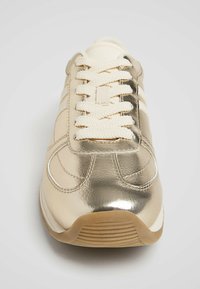 Metalen gouden sneaker met crèmekleurige veters, voorzien van een glad bovenwerk, gebogen ontwerp en een rubberen zool met textuur. Drie strepen aan de zijkant.