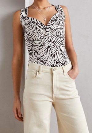 Kobieta ubrana w czarno-białą bluzkę bez rękawów z abstrakcyjnym wzorem i dekoltem w kształcie serca, kremowe wysokowalne dżinsy oraz warstwowe srebrne naszyjniki.