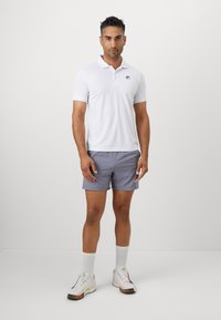Biela polokošeľa s krátkymi rukávmi a malým logom, doplnená svetlošedými šortkami. Outfit dopĺňajú biele velúrové ponožky a atletické topánky.
