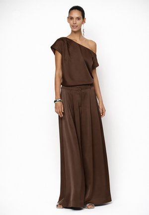 Femme portant un pantalon en satin marron à jambes larges et un haut assorti à une épaule, accessoirisée avec une grande boucle d'oreille et un bracelet argenté, posant devant un fond blanc.